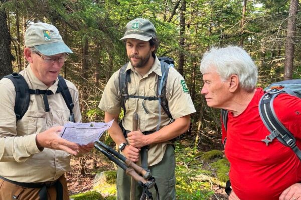 Three men with hiking gear look at a map in a forest.